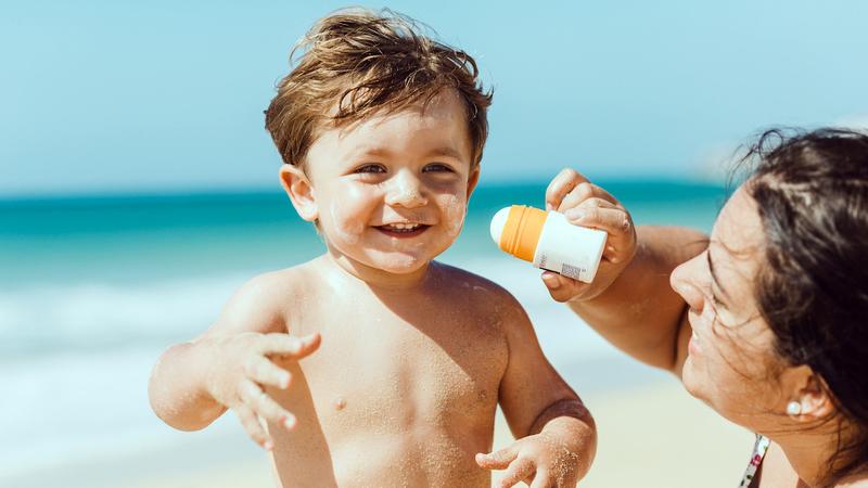 A parent applies sunscreen to a young child at the beach.