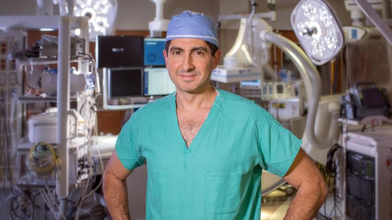 Dr Zayd Eldadah, wearing green scrubs and a blue skull cap, stands with his hands on his hips and looks at the camera in an operating room at MedStar Washington Hospital Center.