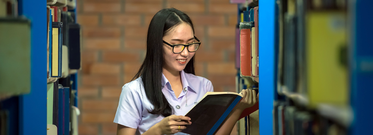 A young woman looks at a book and smiles as while standing between 2 rows of book shelves in a library.