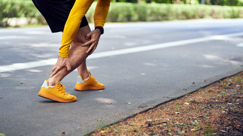 Close up of a man wearing a yellow long-sleeved shirt and yellow shoes grabbing his lower leg in pain during a workout. His face is not visible.