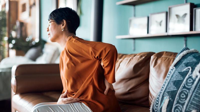 Photo of an older asian woman sitting on a couch in her living room, with her hand on her back, wincing in pain.