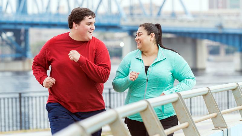 An overweight Hispanic woman and a young mixed race Hispanic and Caucasian man exercising together outdoors in an urban setting, running or jogging along a river with a bridge in the background. They are smiling, looking at each other as they exercise.