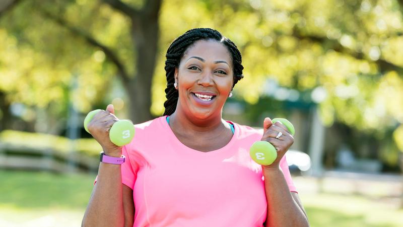 A mid adult African-American woman, in her 30s, with braided hair and large build, wearing a pink t-shirt, exercising in the park, wearing a fitness tracker and lifting 3 pound dumbbells in her hands.