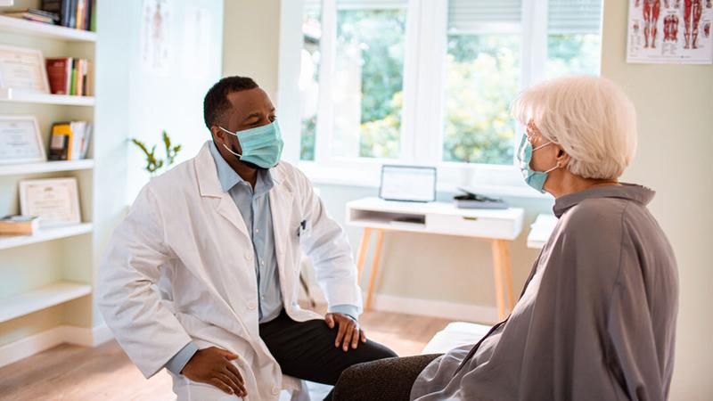 An african american male doctor wearing a masks talks to a senior patient, also wearing a mask, in a clinical setting.