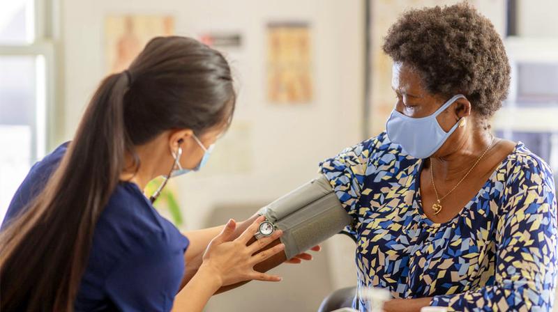 A mature adult female patient has her blood pressure taken by a nurse wearing dark blue scrubs. Both women are wearing masks.