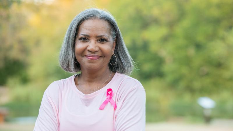 Portrait of a smiling senior mature woman wearing a pink shirt and a pink cancer ribbon, standing outdoors with trees in the background.