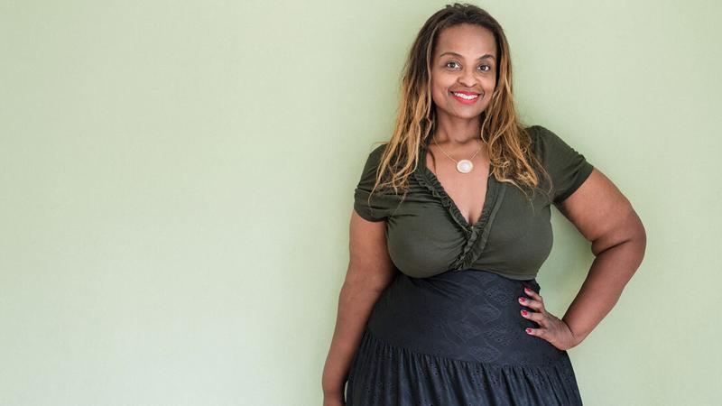 A middle-aged african american woman stands with her hand on her hip againts a plain light green wall.