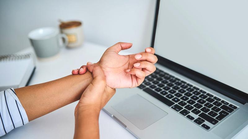 Close up photo of a young adult's wrist as they massage the wrist after too much computer work. A laptop computer and coffee cup are in the background.