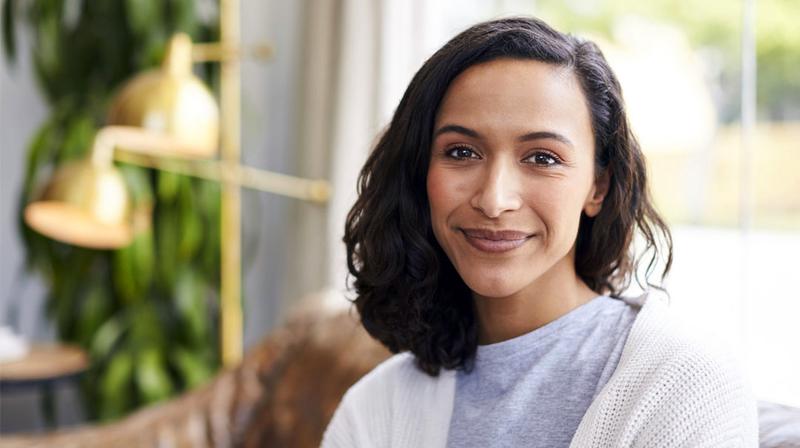 A young woman wearing a white sweater sits in front of a window in her home and smiles slightly as she looks at the camera.