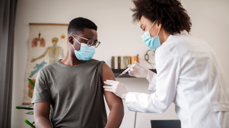 A female doctor gives an african american male patient a vaccination shot in a clinical setting. The patient is looking down at the injection needle as the vaccination is being administered.