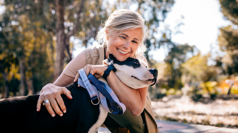 A senior woman smiles as she hugs her large black and white dog outdoors in a park.