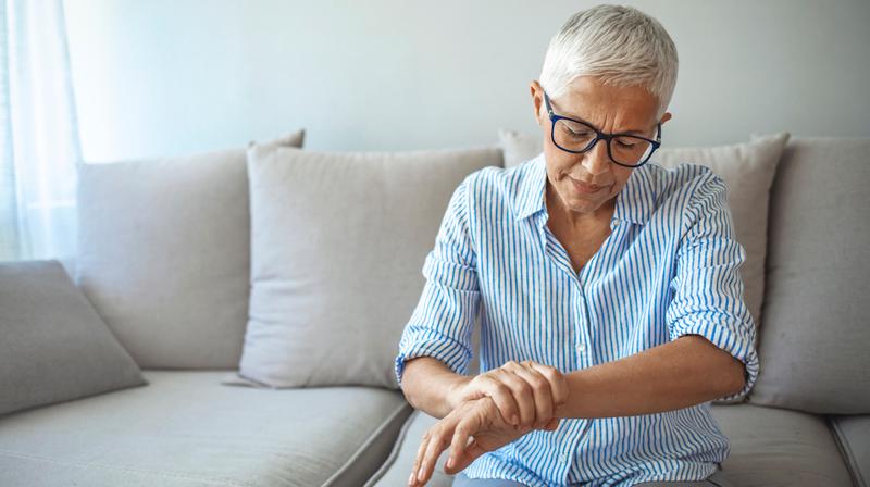 A senior woman looks down at her wrist with concern while sitting on the sofa at home in her livingroom.