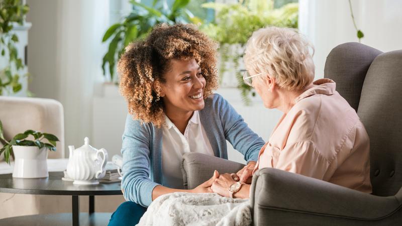 Female home caregiver talking with senior woman, sitting in living room and listening to her carefully.