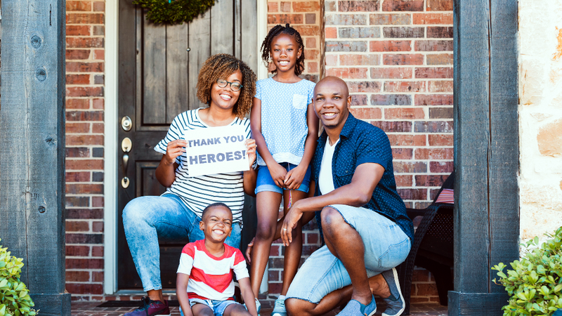 A family of 4 people with two young children pose for a photo in front of their home, and hold a sign that reads "Thank You Heroes."