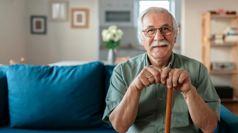 幸福的老人的肖像微笑在家里，而拿着拐杖。成熟的成年男子坐在沙发上，看着相机。