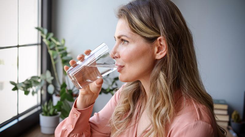 A middle-aged woman stands near a window and drinks a glass of water.