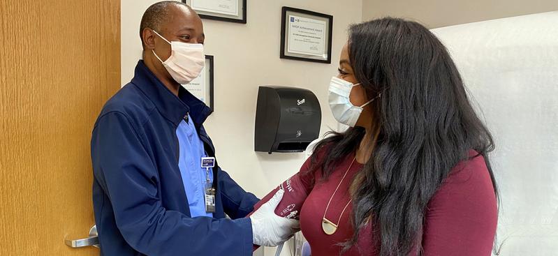 A male nurse takes the blood pressure of a female patient in a clinical setting. Both people are wearing masks.