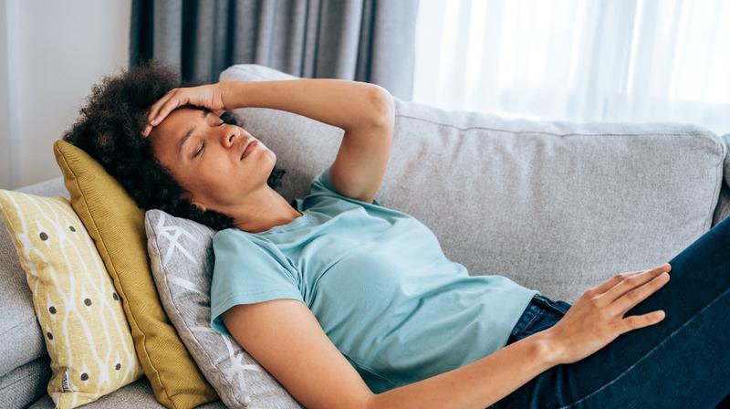 A woman lays on the couch in her living room with her hand on her forehead.