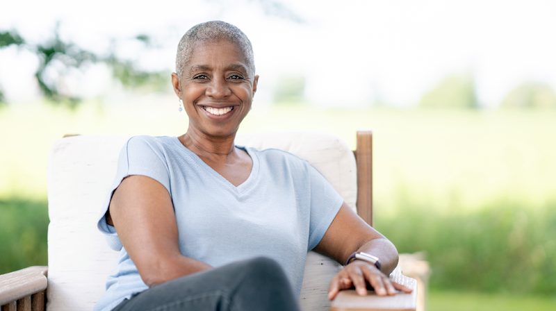 A senior african-american woman sits on a bend outdoors under a tree.