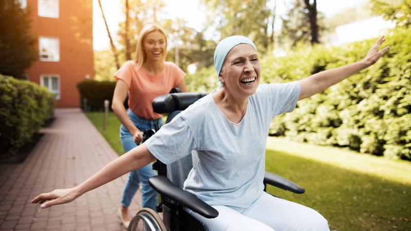 A woman pushes a cancer survivor patient in a wheelchair outdoors.