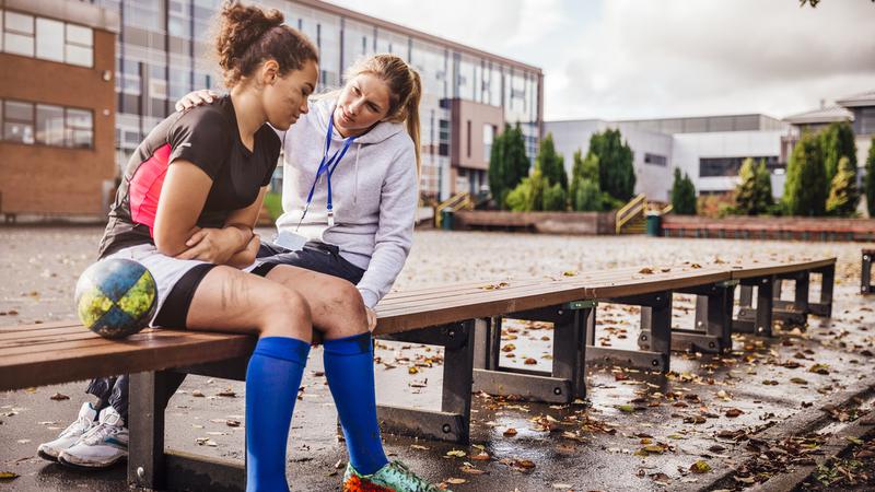 A young soccer player sits on a bench outside of school and is consoled by her female coach.