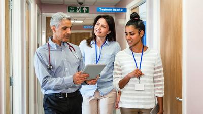 3 healthcare professionals walk down a hallway in a medical office while having a discussion..