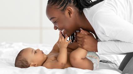 An african-american woman smiles at her infant who is laying on a bed at home.