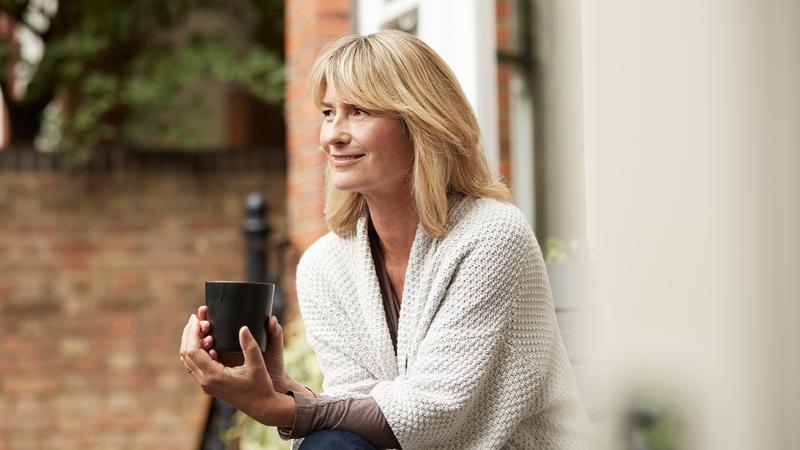 A mature woman holds a cup of coffee while standing outside in her garden.