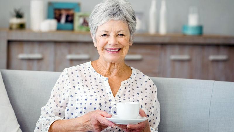 A senior woman sits on her sofa and holds a cup and saucer.