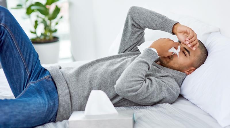 A young african american man lays on a sofa and blows his nose while resting a hand on his forehead.