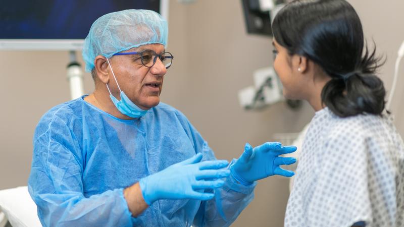 A surgeon, dressed in blue scrubs, talks with a patient prior to performing a procedure.