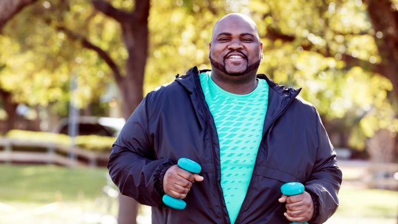 A heavy-set man holds a small dumbbell in each hand as he exercises in an outdoor setting. He is looking at the camera and smiling.