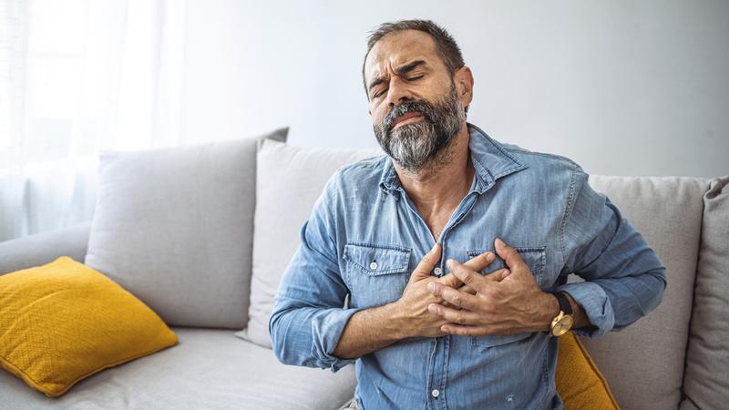 An older man with grey hair sits on his living room sofa and clutches his chest in pain.