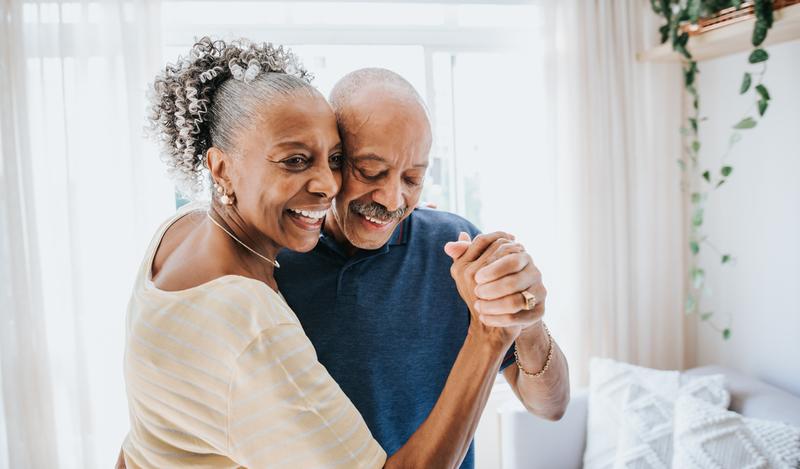 An older African American couple joyfully dances check to cheek in their living room.