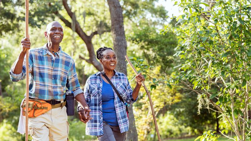 A senior African-American couple in their 70s enjoying the outdoors, hiking in a park. They are walking together with trees and lush foliage in the background, holding hands.