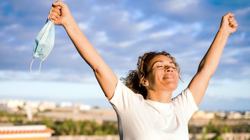 A woman holds her arms up while holding a mask in her hand and smiles outdoors at sunset.