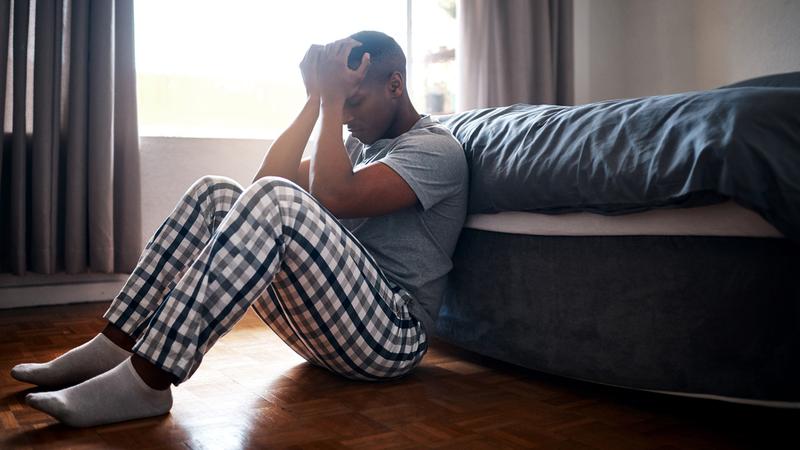 A young man sits on the floor of his darkened bedroom with his head in his hands.