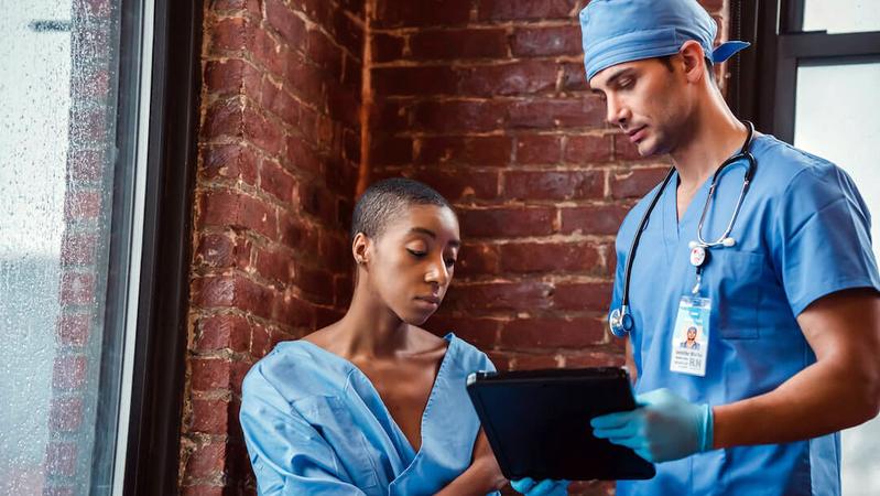 A medical professional wearing blue scrubs, holds a clip board and talks with a patient.