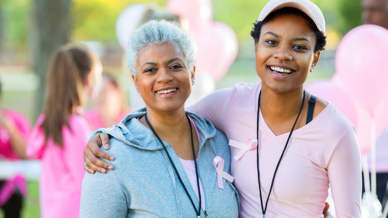 Two women, one older one younger, wearing pink breast cancer support ribbons stand together and smile for the camera at an outdoor event.