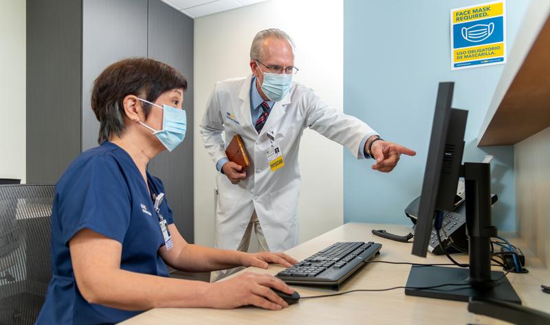 A doctor points to the computer screen while a nurse sits at a desk and types.