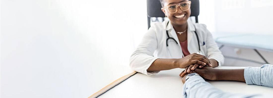 A female doctor, sitting across a table from a patient, places her hands on top of her patient's hands, offering comfort.