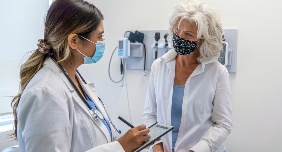 A doctor talks with a mature adult woman in an exam room at a medical office. Both of the women are wearing masks.