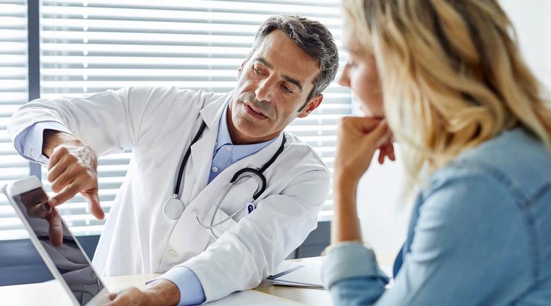 A male doctor points to his ipad while consulting with a female patient in an office setting.