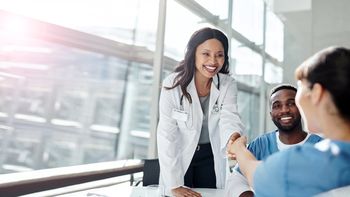 A female doctor shakes hands with another medical professional wearing blue scrubs during a meeting in a brightly lit conference room.