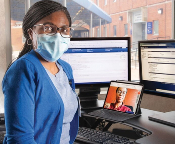 A MedStar Health provider poses for a photo in front of several computers, one of them with a female patient who has called in for a telehealth visit.