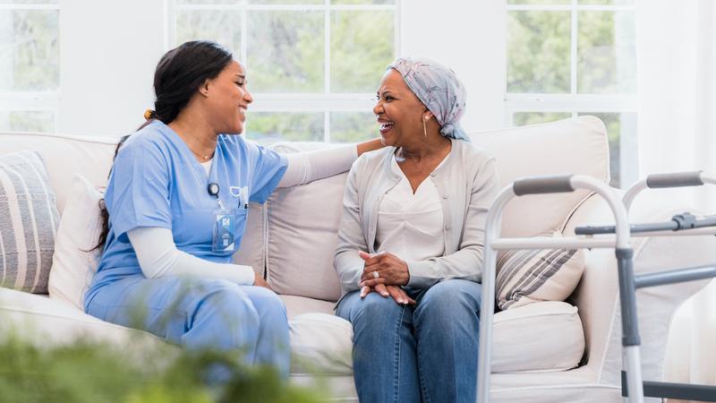 A nurse sits and talks with a patient, who is wearing a head scarf, on a couch.