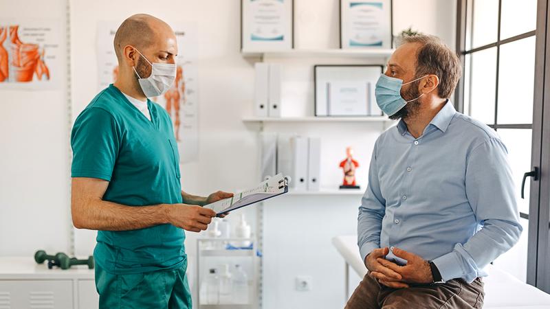 A male doctor wearing green scrubs talks with a male patient in a clinical setting. Both people are wearing masks.