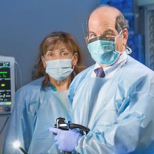 Dr David Shocket holds a colonoscope in a treatment room at MedStar Health. A female physician assistant stands behind him. Both people are wearing blue scrubs and masks.