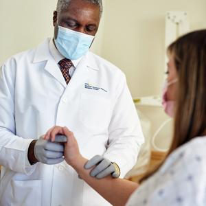 Dr Ahamefula Onyike examines the wrist of a patient in a clincial setting. Both people are wearing masks.