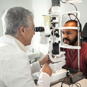 A mature male doctor performs an eye exam on an african american male patient in a clinical setting. The patient has his head in a device which allows the eye doctor to look into his eye.